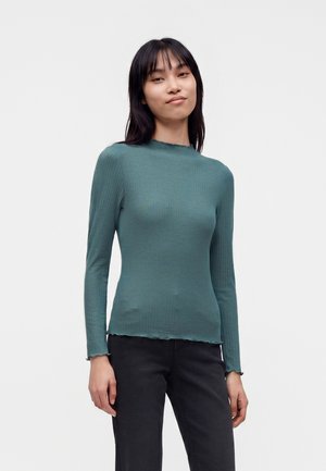 Jeune femme aux cheveux noirs raides portant un haut côtelé bleu sarcelle à manches longues et un pantalon noir, debout devant un fond blanc uni.