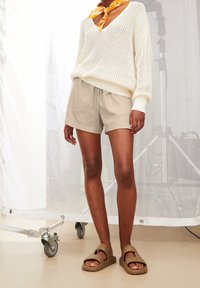 Pull en maille léger de couleur crème, associé à un short beige et des sandales à enfiler marron. Le modèle porte un foulard à motif en guise d'accent.