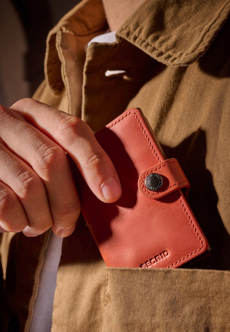 Cartera de cuero roja con cierre de botón, con costuras visibles y un logo. Sostenida en una mano, parcialmente metida en un bolsillo de chaqueta marrón.
