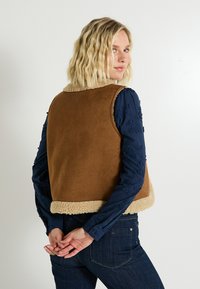 Mujer rubia que lleva un chaleco de borreguillo marrón sobre una camisa de manga larga bordada azul marino, con las manos entrelazadas detrás de su espalda.