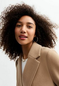 Cappotto in misto lana marrone con rever a punta, indossato su un top di colore chiaro, accessorized con una collana d'oro e orecchini a cerchio.