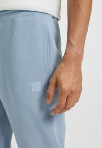 Jasnoniebieskie spodnie dresowe mają gładką teksturę, elastyczny pas, boczną kieszeń oraz kwadratową naszywkę z logo marki na udzie.