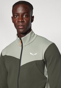 Veste zippée verte et grise avec une surface texturée, présentant un col montant et un logo blanc sur la poitrine, détaillée avec un tirette de zip orange.
