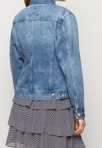 Veste en denim bleu clair, avec un col classique, des boutons comme accents et une texture légèrement délavée. Détails de couture visibles.