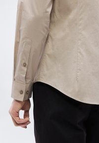 Chemise beige à boutons avec manches longues, présentant une texture lisse, une coupe ajustée et des poignets avec deux boutons arrondis. Pantalon noir visible en dessous.