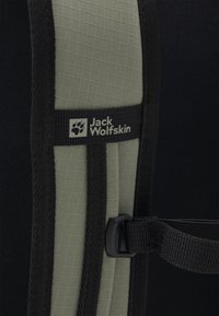Primo piano di una tracolla imbottita verde e nera di uno zaino con il logo Jack Wolfskin e una fibbia nera in plastica regolabile.
