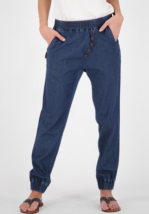 Femme portant un pantalon de jogging bleu décontracté avec détail bouton et poignets élastiques, mains dans les poches, associé à des sandales noires et un haut blanc.