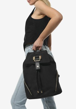 Mochila negra hecha de tela con hardware en tono dorado, tapa superior y dos bolsillos laterales con cremallera. Presenta un diseño elegante y estructurado.
