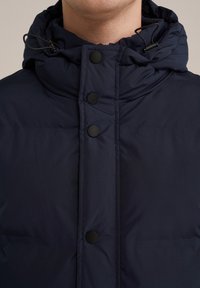 Navy blue puffer jacket featuring a high collar, black snap buttons, and an adjustable drawstring at the neckline for added warmth.