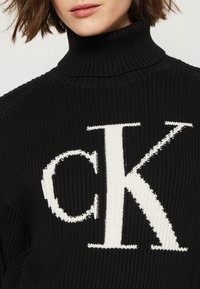 Črn rebrast pulover s turtleneck ovratnikom, ki v sprednjem delu ponuja velik bel logo "ck", s poudarjenim pletenim teksturo in kontrastnimi barvami.