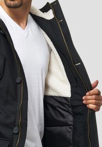 Veste noire avec une fermeture éclair dorée, dotée d'une doublure blanche et duveteuse, ainsi qu'une poche à bouton sur la gauche. Portée ouverte pour révéler le design intérieur.