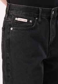 Black denim jeans with a straight cut, featuring a visible Calvin Klein label, metallic hardware, and detailed stitching.