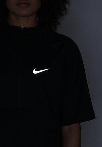 Černý sportovní top s krátkým rukávem, s vysokým límcem a reflexním bílým logem Nike na levé části hrudníku. Hladká textilie.