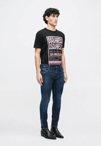 Camiseta de algodón negra con texto gráfico rosa, combinada con jeans skinny azul oscuro y botines de cuero negros. El modelo está de pie frente a un fondo liso.