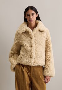 Femme portant une veste en shearling beige fluffy à boutons avec un large col et un pantalon en velours côtelé marron ample, se tenant devant un fond uni.