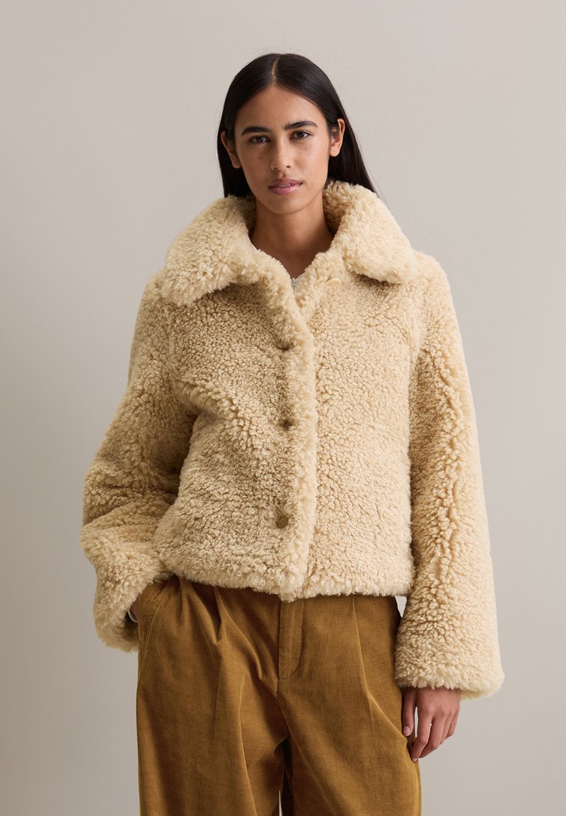 Femme portant une veste en shearling beige fluffy à boutons avec un large col et un pantalon en velours côtelé marron ample, se tenant devant un fond uni.