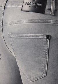 Zbliżenie na tylnią kieszeń jasnoszarych dżinsów z czarną skórzaną metką marki Próchnik i małym czarnym metalowym emblematem.