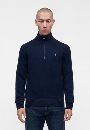 Homme portant un pull en maille côtelée bleu marine avec une fermeture éclair au quart et un petit logo blanc brodé sur la poitrine gauche, associé à un jean foncé.