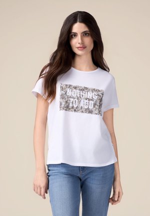 Camiseta blanca de algodón con mangas cortas y corte holgado, que presenta un parche de lentejuelas plateadas con el texto "NADA QUE AÑADIR."