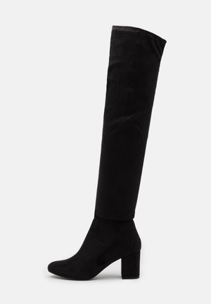 Botte noire en daim montant jusqu’au genou avec un bout rond et un talon bloc, présentée sur un fond clair uni.