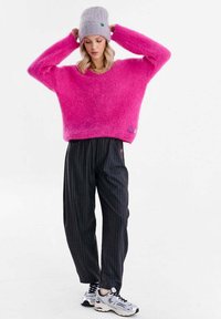 Pulover roz pufoasă, oversized, cu decolteu în V, asortat cu pantaloni negri cu dungi subțiri și o căciulă gri. Tenisii completează ținuta.