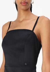 Robe noire à fines bretelles, avec un décolleté carré et un tissu lisse. Comprend un petit logo près de l'ourlet.