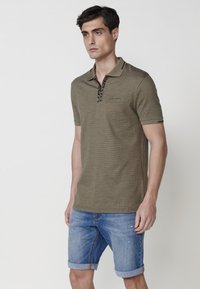 Polo de color verde oliva con mangas cortas, tejido texturizado y un diseño de cuello negro; combinado con pantalones cortos de mezclilla arremangados.