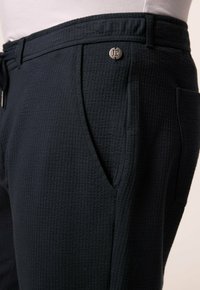 Navy textured pants with a waffle weave, featuring side pockets and a metal button with "JP" logo. Elastic waistband for comfort.