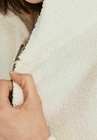 Veste blanche texturée en tissu doux, avec une fermeture éclair argentée et un col confortable. Une main tient le bord près de la fermeture éclair.