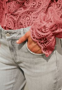 Roza čipkasta bluza z zapletenimi vzorci, kombinirana s svetlo sivimi kavbojkami z žepom za kovance. Bluza ima nabrane manšete in podrobne teksture.