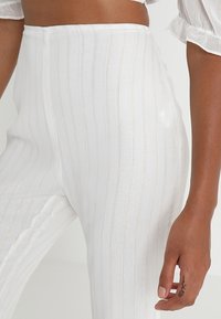 Białe spodnie z wysokim stanem w pionowe paski, z delikatnym połyskiem i teksturowanym wykończeniem. Paski są uzupełnione metalicznymi nitkami.