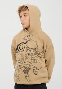 Felpa beige con un disegno a tratto nero di due personaggi, con un simbolo a spirale dietro di loro. Tessuto morbido, maniche lunghe e cappuccio.