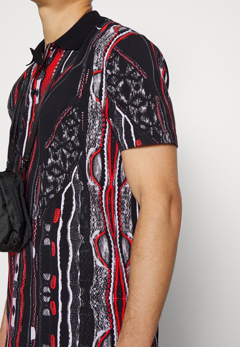 Homme portant un polo noir à manches courtes avec un motif abstrait rouge, blanc et gris, portant un petit sac bandoulière noir.