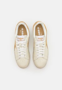 Sneakers made of cream leather with tan suede accents, featuring white laces and a beige sole. Logo displayed on the insole and tongue.