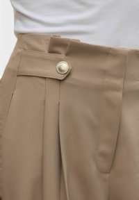 Beige bukser med flad front, to dybe folder og sølvknaplukning i taljen med bæltesløjfer. Glat stofstruktur.
