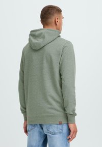 Sudadera verde de tejido suave, con capucha ajustable, mangas largas y dobladillo acanalado. Combinada con jeans de color azul claro.