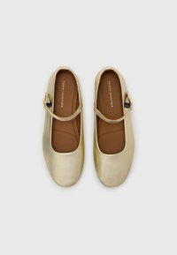 Pantofi în stil Mary Jane din piele aurie, cu vârf rotund, talpă plată și o singură curea peste laba piciorului, având o căptușeală interioară maro.