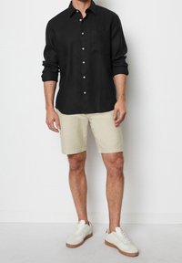 Camisa de lino negra de manga larga con cuello abotonado, botones blancos y un bolsillo en el pecho, combinada con pantalones cortos beis y zapatillas blancas.