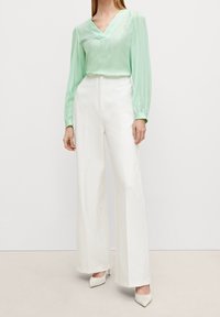 Blusa verde menta a maniche lunghe con scollatura a V; abbinata a pantaloni bianchi a gamba larga e tacchi di colore chiaro per un look coordinato.
