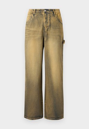 Ξεθωριασμένο κίτρινο-καφέ τζιν με φαρδιά μπατζάκια, μπροστινές τσέπες, κούμπωμα με κουμπί, θηλιές για ζώνη και λεπτομέρεια με πλαϊνή θηλιά σε απλό φόντο.