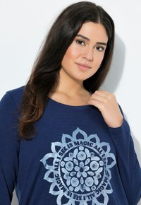 Chemise à manches longues marine en tissu doux, avec un grand motif graphique argenté représentant un design de mandala floral, accompagné de texte disposé en cercle.