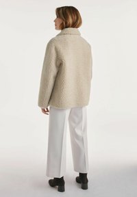 Kobieta zwrócona w lewo, ubrana w beżową, teksturowaną kurtkę polarową, białe szerokie spodnie i czarne botki na obcasie na białym tle.