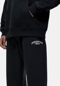Zwarte sweatshirt met een kangoeroezak en zwarte sweatpants met een witte "GOODFORNOTHING Atelier" logo op de bovenbeen. Zachte textuur.