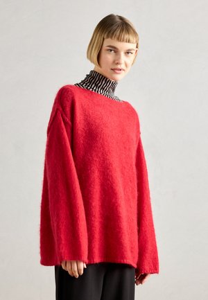 Roter Wollpullover mit lockerer Passform, weiten Ärmeln und einem runden Ausschnitt, über einem schwarz-weißen gestreiften Rollkragenpullover getragen.