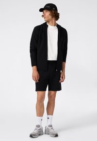 Sudadera negra con cremallera, camiseta blanca, pantalones cortos negros, zapatillas grises y una gorra negra, con detalles de logotipo y texturas que contrastan.