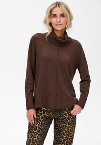 Kobieta z blond włosami, ubrana w brązowy sweter z golfem i spodnie w leopardzie cętki, stojąca na tle białym.