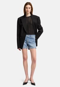 Blazer oversize nero, top trasparente con motivo, pantaloncini in denim blu chiaro con orlo sfrangiato e décolleté neri a punta. Design minimalista.