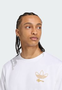 Witte katoenen T-shirt met een gouden geborduurd logo met een driebladig ontwerp en het woord "Tigres" eronder.