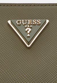 Grön triangulär Guess USA-logotyp med guldtext och frågetecken på en strukturerad olivgrön handväskayta.