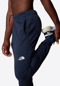 Mörkblå joggers med en texturerad tyg, elastisk midja och en synlig logotyp på låret. Höger ben är böjt, vilket visar passformen.